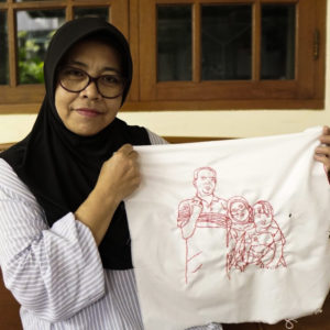 A picture of Ibu Dwi Restu smiling and holding up her final embroidery of her family that features three figures. She is sitting on a couch in front of a wood framed window.