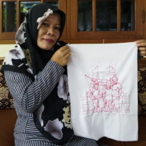 Ibu Nisa is holding up her final embroidery of her family. She is smiling into the camera. She is wearing a black flowered hijab and long striped sleeves. She is standing in front of wooden cabinets with glass panels.