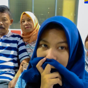 Picture of Ibu Dwi Restu's and her husband and daughter. Her daughter is looking into the camera, holding her hand to her face covering her mouth with her hijab in hand, in the foreground wearing a blue hijab and her husband is in the background looking at the camera with a blue and white striped shirt. Ibu Dwi Restu is wearing an orange hijab and is looking into the camera.