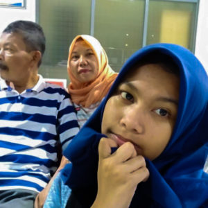 Picture of Ibu Dwi Restu's and her husband and daughter. Her daughter is looking into the camera holding her hand to her face in the foreground wearing a blue hijab and her husband is in the background looking away with a blue and white striped shirt. Ibu Dwi Restu is wearing an orange hijab and is looking into the camera.