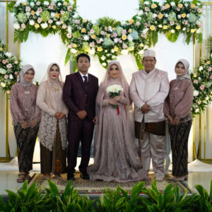 A picture of Ibu Dwi Restu's wedding. Six people are standing until a garland of greenery and baubles. Ibu Dwi is in the middle holding a bouquet of white flowers and is wearing a light purple gown. Her husband is standing next to her. Everyone is smiling and dressed beautifully.