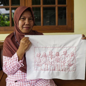 A picture of Ibu Dwi Restu smiling and holding up her final embroidery of her family. It features six figures in a rectangle. She is sitting on a couch in front of a wood framed window.