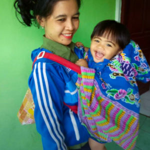 A picture of Ibu Yuli smiling with her young son in a baby carrier with a red strap, smiling in front of her. They are in front of a bright green wall and Ibu Yuli is wearing a bright blue track suit.