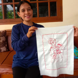 A picture of Ibu Yuli smiling and holding up her final embroidery of her family. It features four figures in a rectangle. She is sitting on a couch in front of a wood framed window.