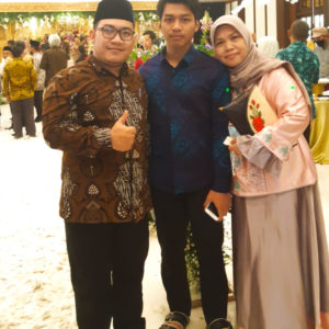A picture of Ibu Quodar and her family. They are standing in a large room. Ibu Qodar is wearing a pink and grey dress with a grey hijab. She is leaning in close next to her son who is wearing a blue shirt and black pants. Her other son is wearing an indonesian hat and a brown patterned shirt with black pants and is giving the thumbs up sign.
