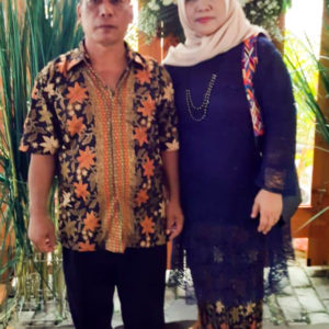 A picture of Ibu Quodar and her husband standing in front of some green trees. Her husband is wearing a flower patterned shirt and Ibu Quodar is wearing a blue dress and a beige hijab.