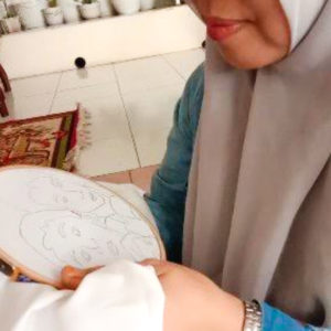 Ibu Quodar is embroidering her piece on an embroidery hoop. She is wearing a beige hijab and a blue longsleeve shirt.
