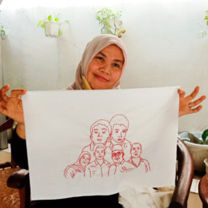 Ibu Munah is in a room with a white wall and some plants and is smiling with a beige hijab on and is holding up her final embroidery of her family. It features six figures.