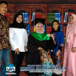 A picture of Ibu Munah and her family. There are five figures all smiling at the camera. Ibu Munah is smiling, wearing a head to toe pink dress with a pink hijab. Next to her is a daughter wearing a bright blue outfit. In the middle is their daughter is sitting and holding up a green medal and is wearing a black graduation cap, she is smiling very big. Next to her stands another daughter wearing a black hijab and a white silk shirt. Next to her is Ibu Munah's husband smiling wearing a patterned shirt and jeans.