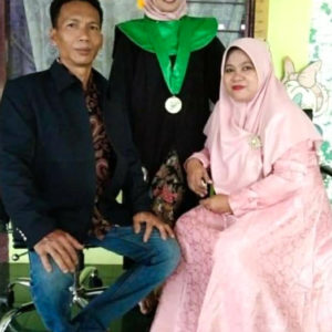 A picture of Ibu Munah and her family. She is sitting and smiling, wearing a head to toe pink dress with a pink hijab. Her husband is sitting next to her smiling wearing a black jacket and jeans. Their daughter is between them wearing a green medal and black graduation cap, she is smiling very big.