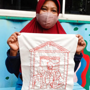 Ibu Munah is standing in front of a turquoise background and is smiling with a mask and red hijab on and is holding up her final embroidery of her family. It features three figures standing in front of a building with a peaked roof.