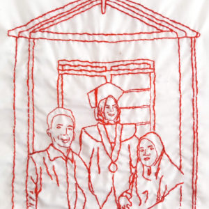 Ibu Munah's final embroidery of her family. It features three figures standing in front of a building with a peaked roof. The girl in the middle has a graduation cap on and is smiling.