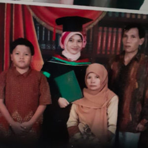 A picture of Ibu Suyanah's family. It features four figures standing together. The middle figure has a graduation cap on and is holding a diploma. She looks happy. She is surrounded by a young boy and a young girl and a man.
