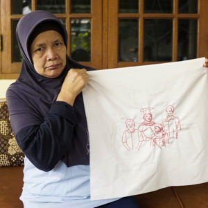 Ibu Suyanah is holding up her final embroidery of her family. She is holding it up and staring into the camera. She is wearing a navy blue hijab and long sleeves with a white shirt. She is standing in front of wooden cabinets with glass panels.