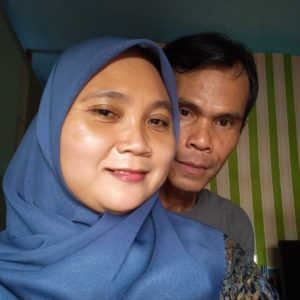 A picture of Ibu Dahlia and her husband from the shoulders up. She is wearing a blue hijab and is smiling in front of her husband. He is behind her staring into the camera.