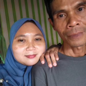 A picture of Ibu Dahlia and her husband from the shoulders up. She is wearing a blue hijab and is smiling. Her hand is on her husbands shoulder. They look happy and peaceful.