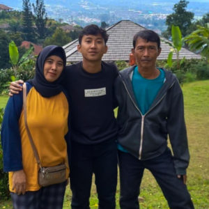 A picture of Ibu Mariah, her son and husband standing together. Her son is in the middle with his arm around her. She is wearing a yellow shirt with a blue hijab, her son is wearing all black and her husband is wearing a grey sweatshirt with a turquoise shirt underneath. They are standing in a green grass area with mountains and a house behind them.