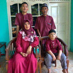 A picture of Ibu Mariah's family. She is sitting on a chair with her youngest son sitting next to her on a chair. Behind them are her older song and husband standing. They are all wearing red and are in front of a turquoise wall with a white door and cabinet.