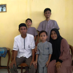 A picture of Ibu Marini's family. There are three boys of different ages. Ibu Marini is seated in the front and is wearing a hijab, her husband is wearing a white shirt and an indonesian hat. They are all in front of a sunny yellow wall.